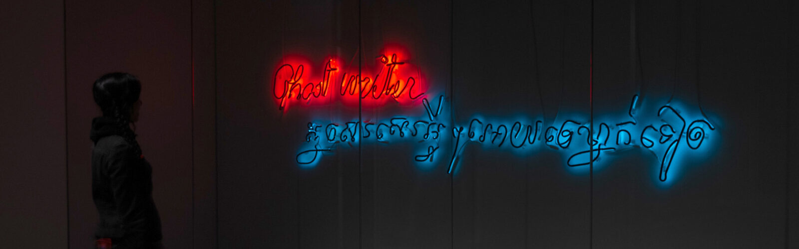 A figure standing in front of neon sign that reads "Ghostwriter" in English and Khmer